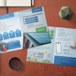 Ein aufgeschlagener Flyer auf einem Schreibtisch informiert auf Deutsch über eine Solaranlage. Er zeigt Bilder von Solarmodulen, einer Batterie, einem Haus und einer Smartphone-App. Daneben liegen ein Stift, ein geometrisches Objekt und ein Behälter mit Büroklammern.