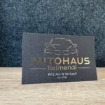 Eine schwarze Visitenkarte mit goldenem Text und einer Auto-Strichzeichnung. Darauf steht: „AUTOHAUS Kelmendi, KFZ An- & Verkauf, seit 1995.“ Die Karte steht aufrecht auf einer Holzfläche vor dunklem Hintergrund.