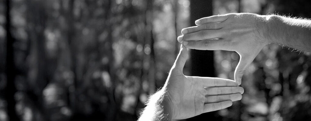 Zwei Hände, die eine rechteckige Form vor einem unscharfen Hintergrund im Freien einrahmen, suggerieren den Akt der Visualisierung oder Planung einer Szene. Das Bild ist in schwarz-weiß gehalten.