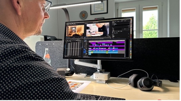 Eine Person sitzt an einem Schreibtisch und arbeitet mit einer Videobearbeitungssoftware auf einem großen Monitor; daneben liegen Kopfhörer, eine Wasserflasche und andere Büromaterialien.