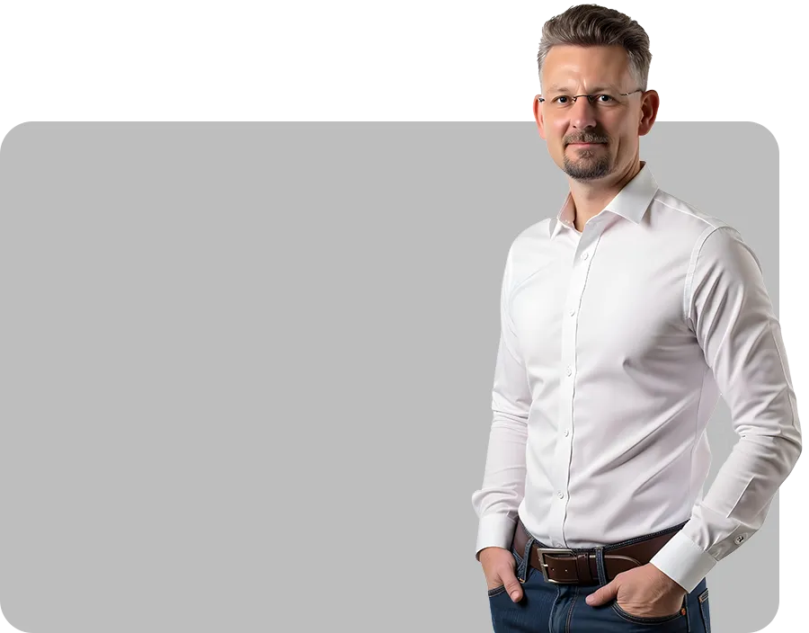 Vor einem grauen Hintergrund steht ein Mann mit Brille und gepflegtem Bart, der ein weißes Hemd und blaue Jeans trägt und lächelnd die Hände in den Taschen hat.