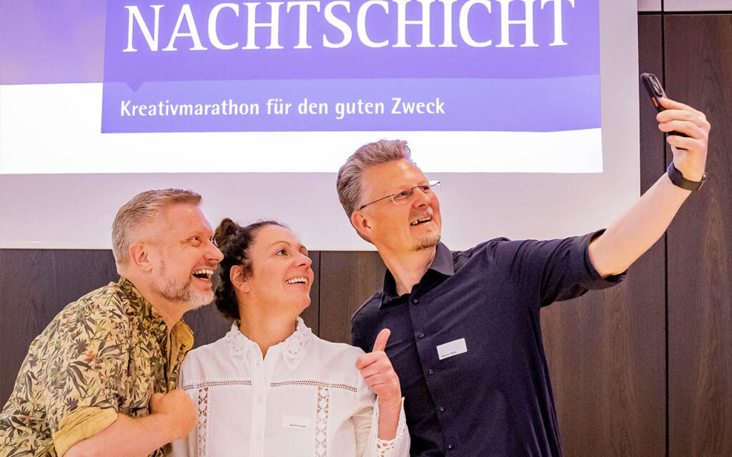 Drei Menschen, die lächeln und gemeinsam in einem Raum ein Selfie machen, während sie vor einem Schild stehen, auf dem „NACHTSCHICHT Kreativmarathon für den guten Zweck“ steht.