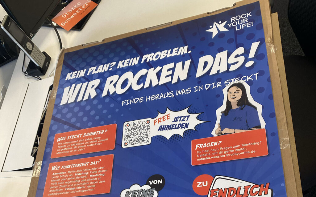 Auf einem Schreibtisch steht ein blaues Plakat mit der Aufschrift „Wir rocken das!“, darauf sind eine Comic-Figur, Sprechblasen und ein „Rock Your Life!“-Logo zu sehen. An den Rändern sind teilweise weitere Bürogegenstände zu erkennen.