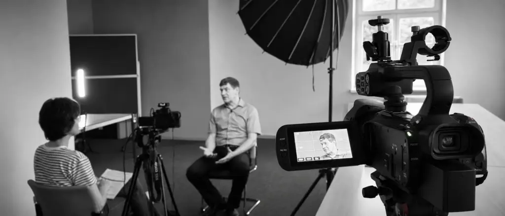 Ein Mann wird in einem Studio vor der Kamera interviewt. Ihm gegenüber sitzt eine weitere Person mit Unterlagen, während um sie herum Aufnahmegeräte und Scheinwerfer aufgestellt sind. Die Szene ist in Schwarz-Weiß gehalten.