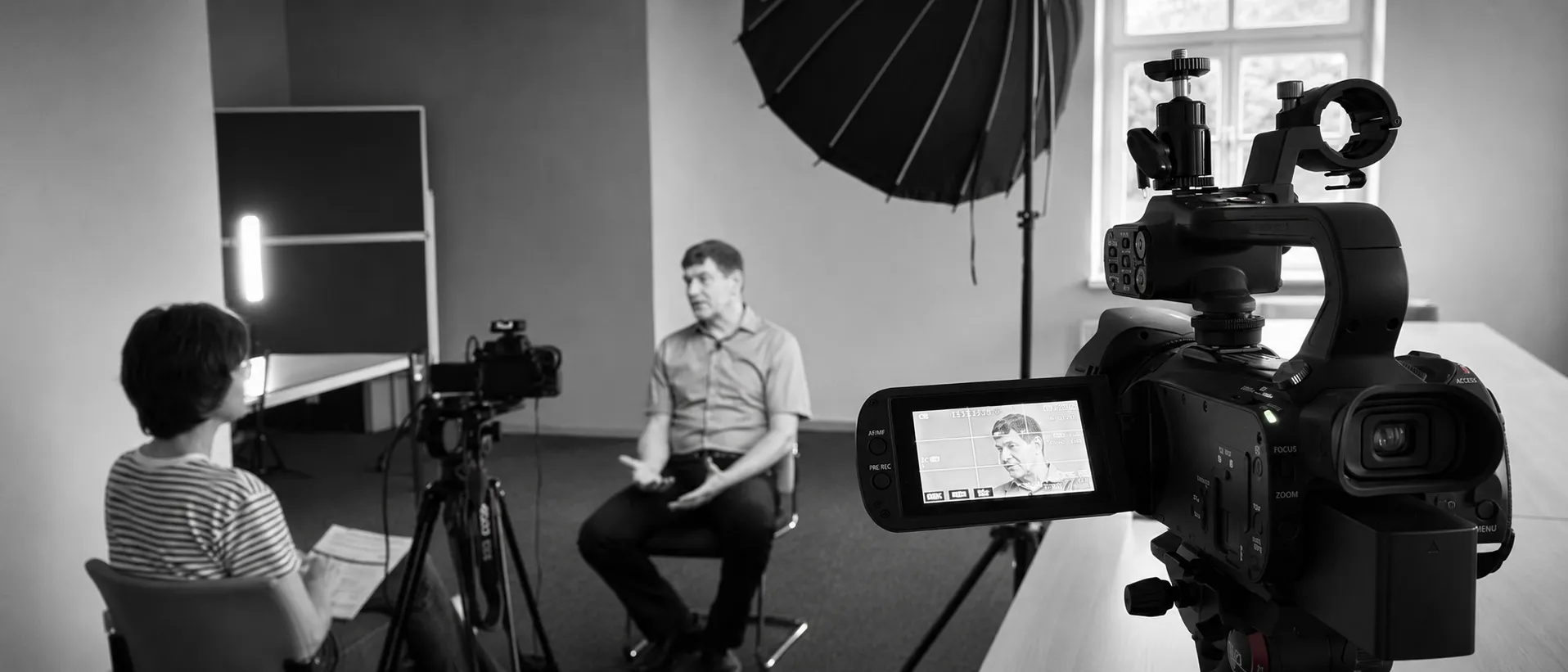 Ein Mann wird in einem Studio vor der Kamera interviewt. Ihm gegenüber sitzt eine weitere Person mit Unterlagen, während um sie herum Aufnahmegeräte und Scheinwerfer aufgestellt sind. Die Szene ist in Schwarz-Weiß gehalten.