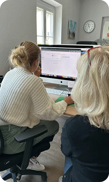 Zwei Frauen sitzen an einem Schreibtisch vor einem Computerbildschirm und arbeiten gemeinsam in einem hellen Büro mit Fenstern und einer Uhr an der Wand.