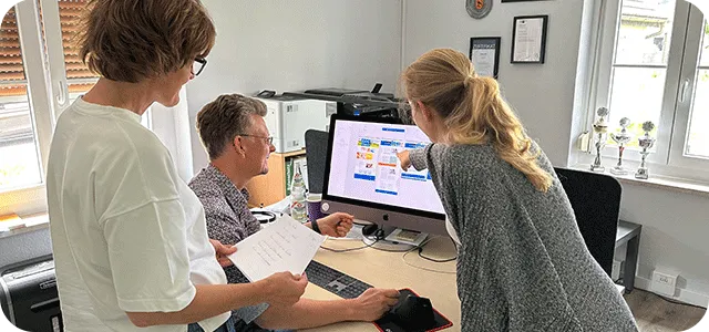 Drei Personen stehen um einen Schreibtisch herum und schauen auf einen Computerbildschirm. Eine Person sitzt am Schreibtisch, während die anderen beiden daneben stehen; eine von ihnen zeigt auf den Monitor. Auf dem Bild sind Papiere und Büromaterial zu sehen.
