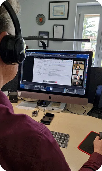 Eine Person mit Kopfhörern sitzt an einem Schreibtisch und nutzt einen iMac für eine Videokonferenz. Auf dem Bildschirm sind mehrere Personen zu sehen. Auf dem Schreibtisch liegen außerdem ein Smartphone und ein Notizblock.