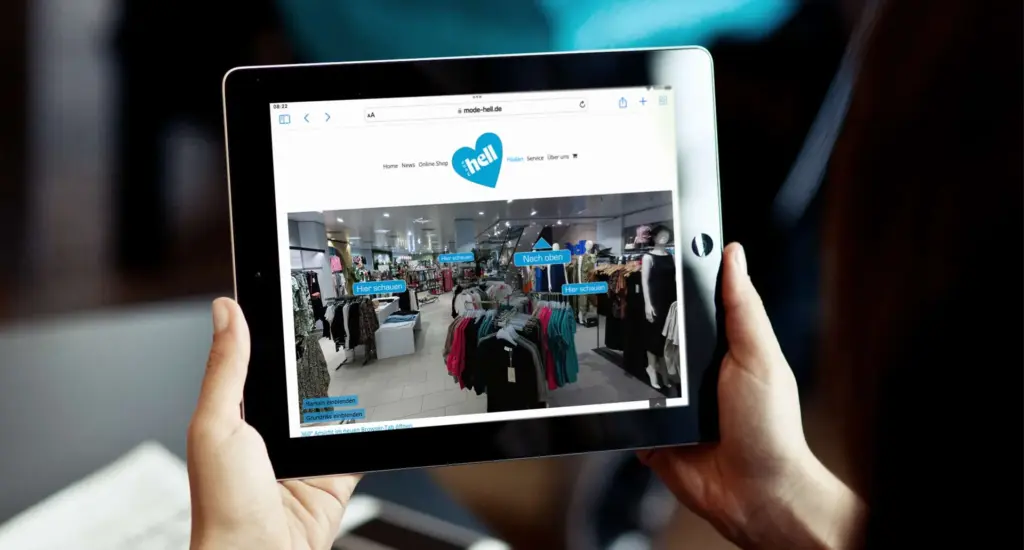 Eine Person hält ein Tablet in der Hand, auf dem ein Online-Kleidershop angezeigt wird. Auf dem Bildschirm sind verschiedene Kleiderständer und Bereiche mit deutschen Beschriftungen zu sehen, darunter „Hier shoppen!“ und „Nach oben“, inmitten eines Ladenlayouts.